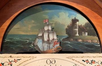 彩色照片显示在一个高表壳时钟的表盘上方的一个新月形的画海景. 这幅画描绘的是一座石砌建筑，矗立在长满草的露头上，海岸线上有岩石. 前面的风景是一艘船(三桅护卫舰).